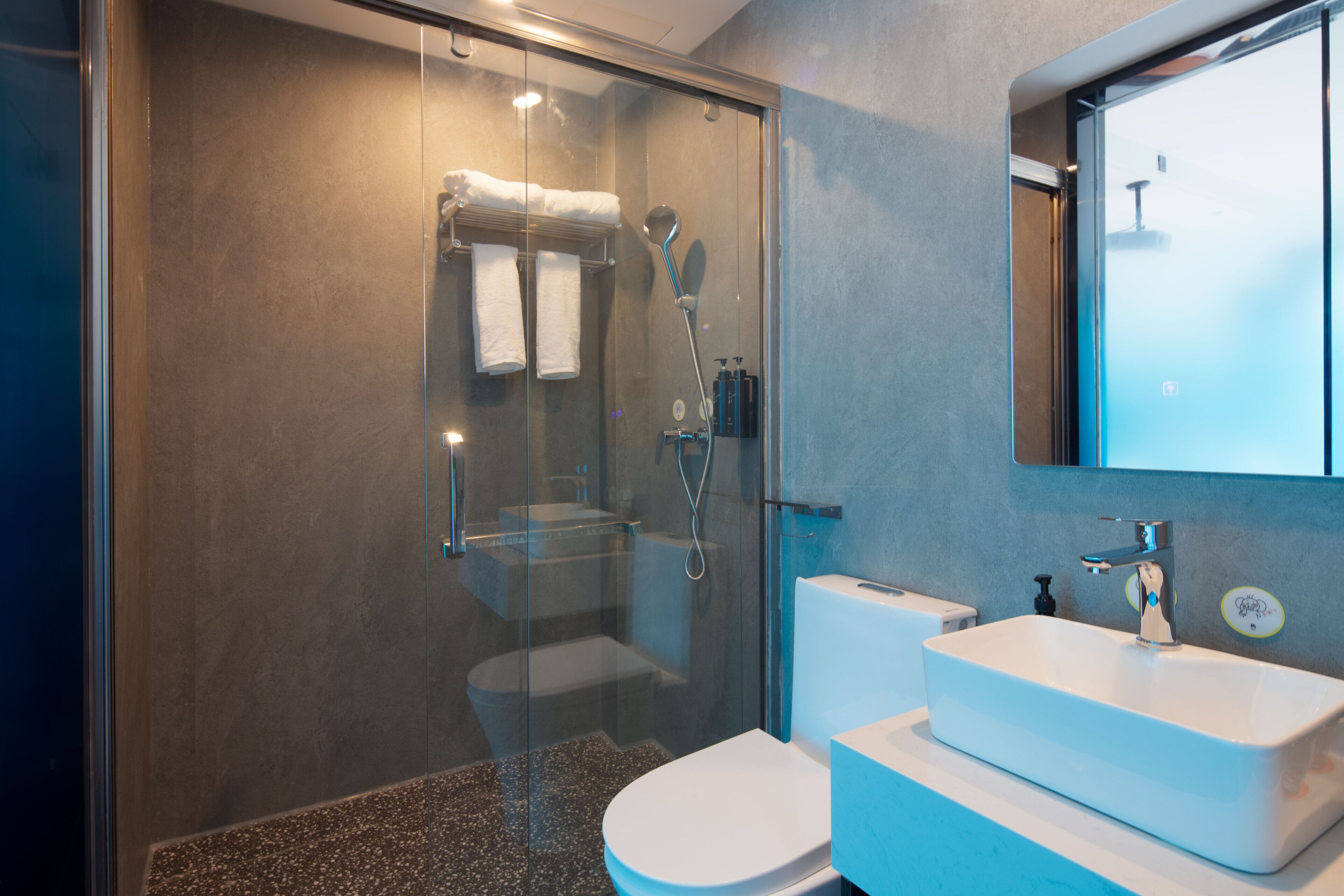 Une salle de bain moderne avec une douche à parois de verre, des toilettes blanches et un grand lavabo rectangulaire surmonté d’un miroir.