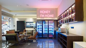 Un moderno vestíbulo de hotel con un letrero de neón que dice "HONEY I'M HOME", áreas de descanso, un refrigerador para bebidas y una exhibición de zapatos.