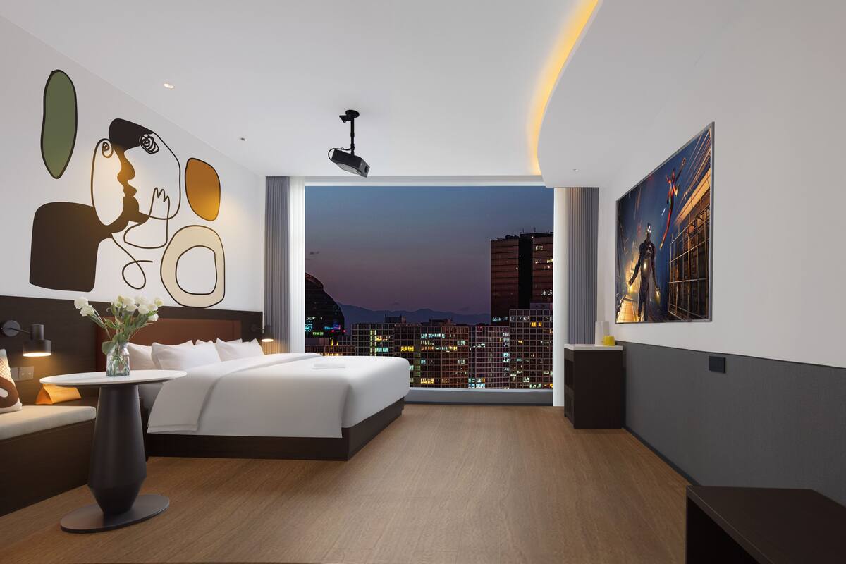 Een moderne hotelkamer met een groot bed, uitzicht over de stad, een projecteerder en een schilderij aan de muur.