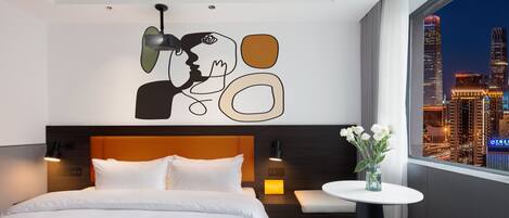 A modern hotel room with a large bed, a bedside table with a vase of flowers, a cityscape view, and a unique abstract wall art piece.