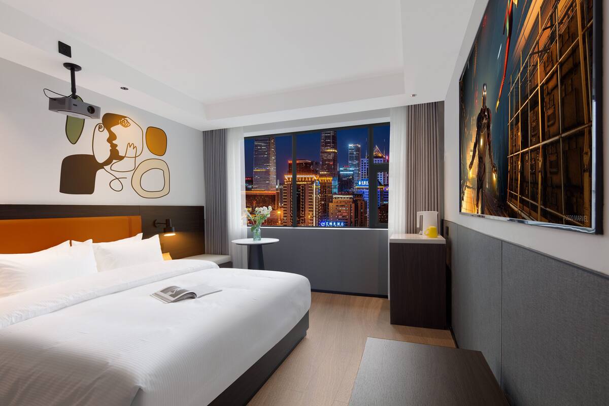 Een moderne hotelkamer met een groot bed, een klein tafeltje met een vaas, een nachtkastje en een stadszicht door het raam.