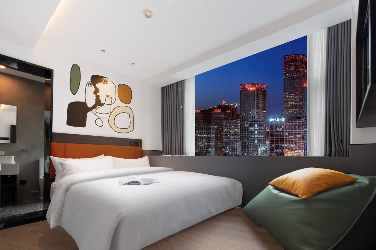 Een moderne hotelkamer met een groot bed, een groene fauteuil en uitzicht op de stad in de schemering.