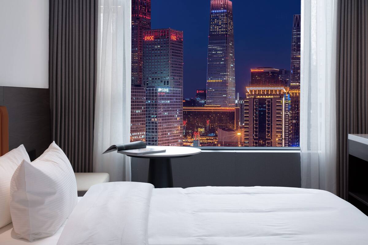 Een hotelkamer met een bed, een klein tafeltje en uitzicht op een stadszicht bij nacht.