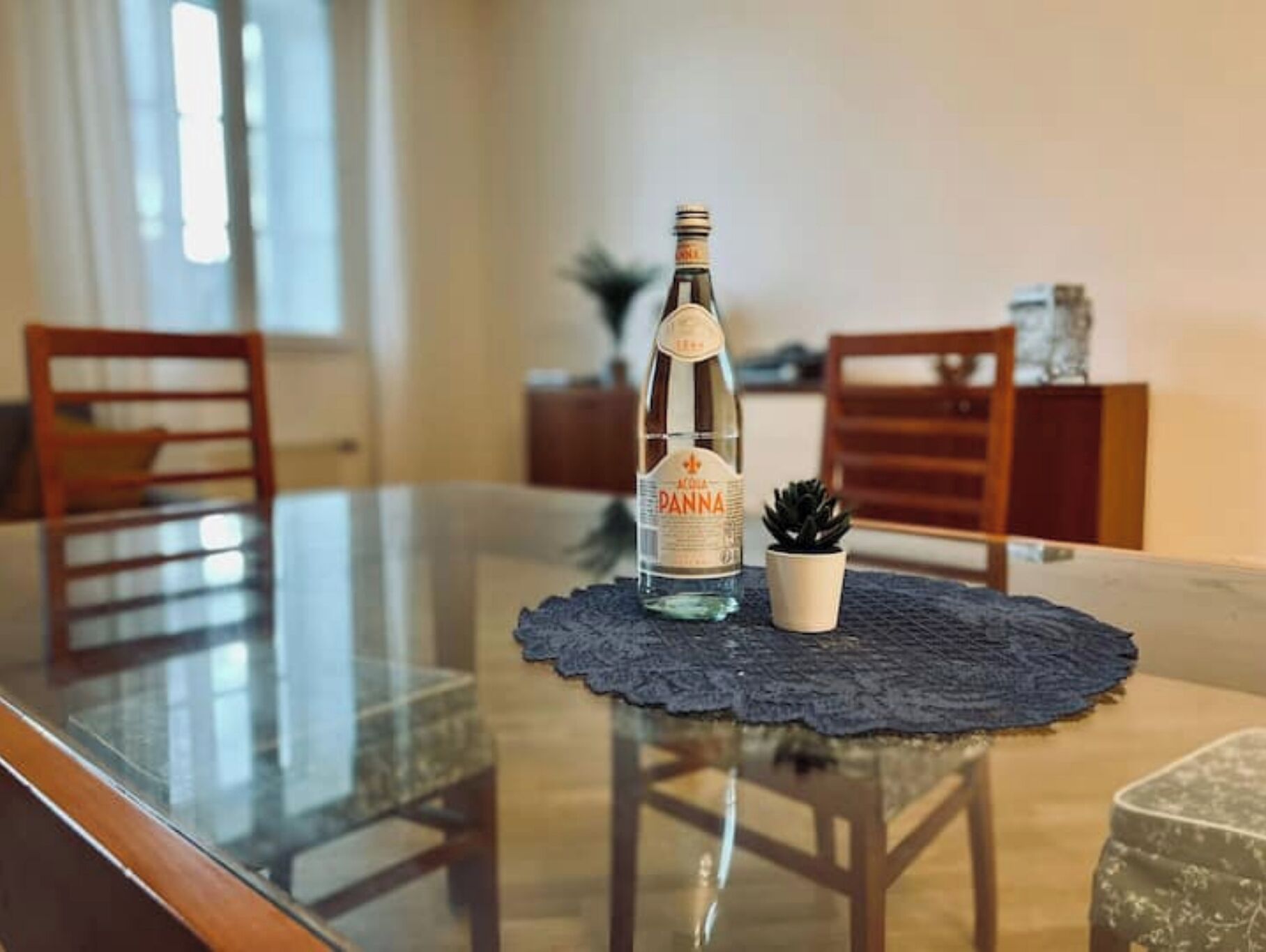 Une table à manger avec une bouteille d’eau Panna, une petite plante en pot et un set de table en dentelle.