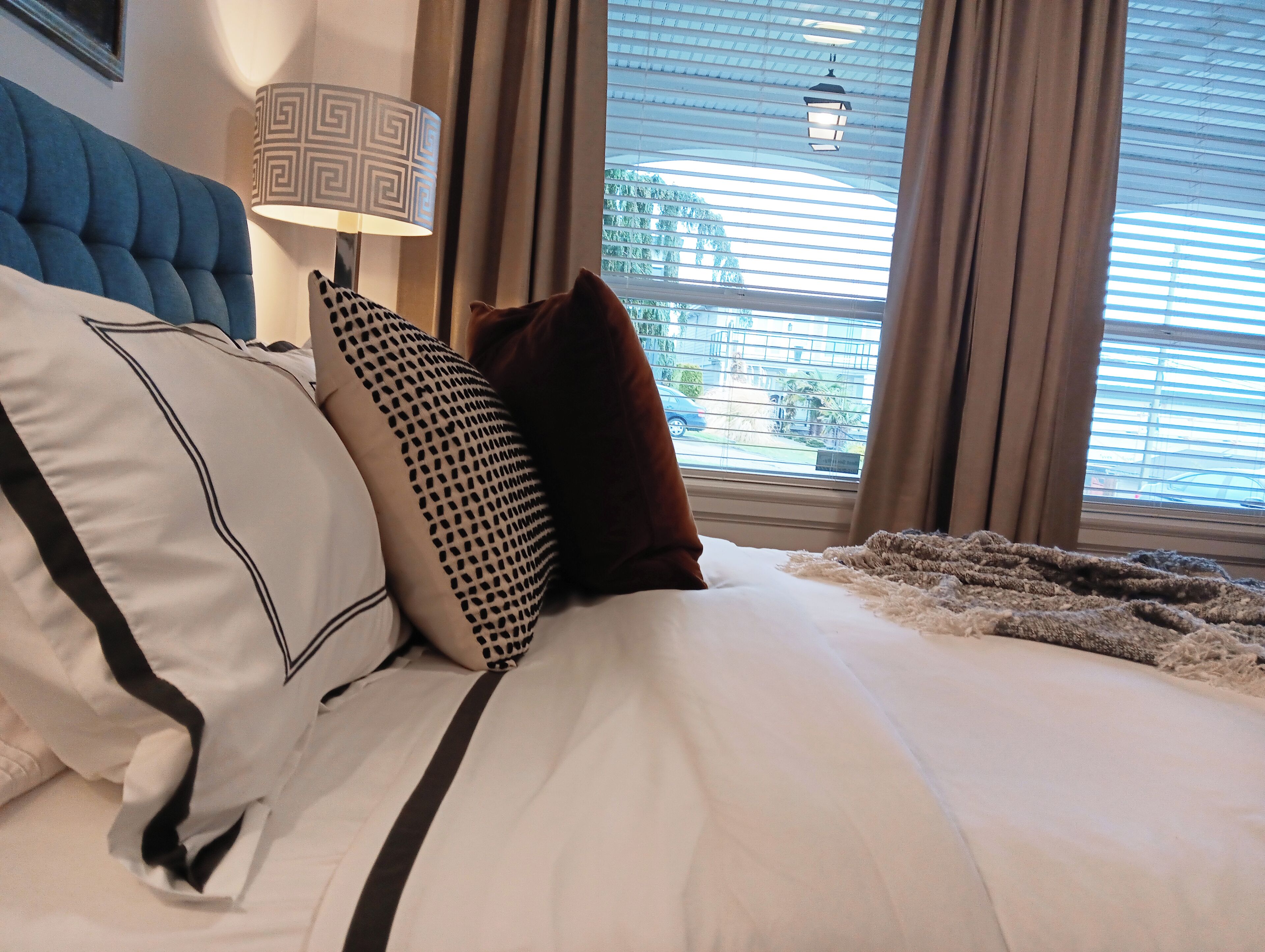 Una cama bien hecha con ropa blanca y detalles negros, una almohada decorativa estampada y un cabecero tapizado en azul.