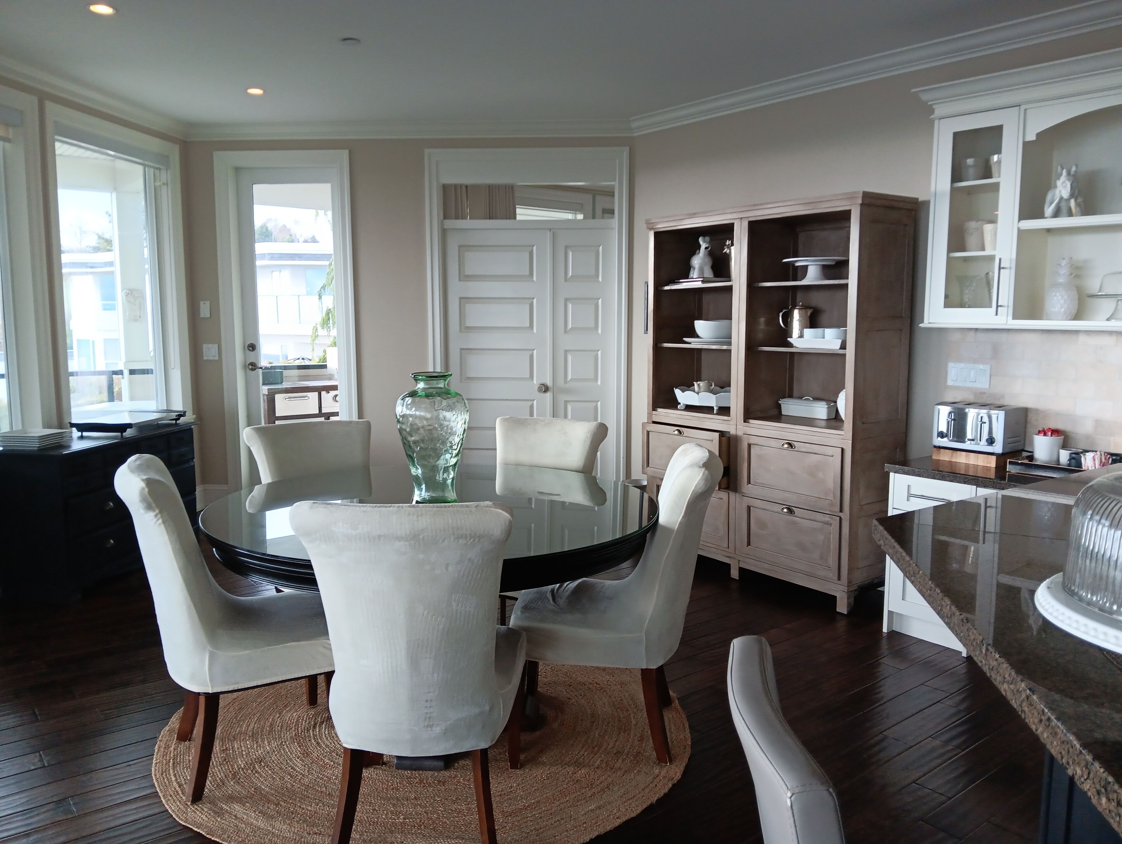 A dining area with a round table, white chairs, and a green vase. There is a kitchen area with cabinets and a toaster.