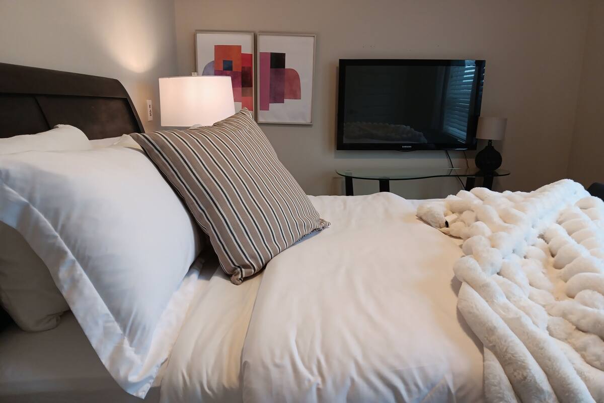 Una cama bien hecha con sábanas blancas y una manta suave, un cojín a rayas y una lámpara en una mesita de noche.