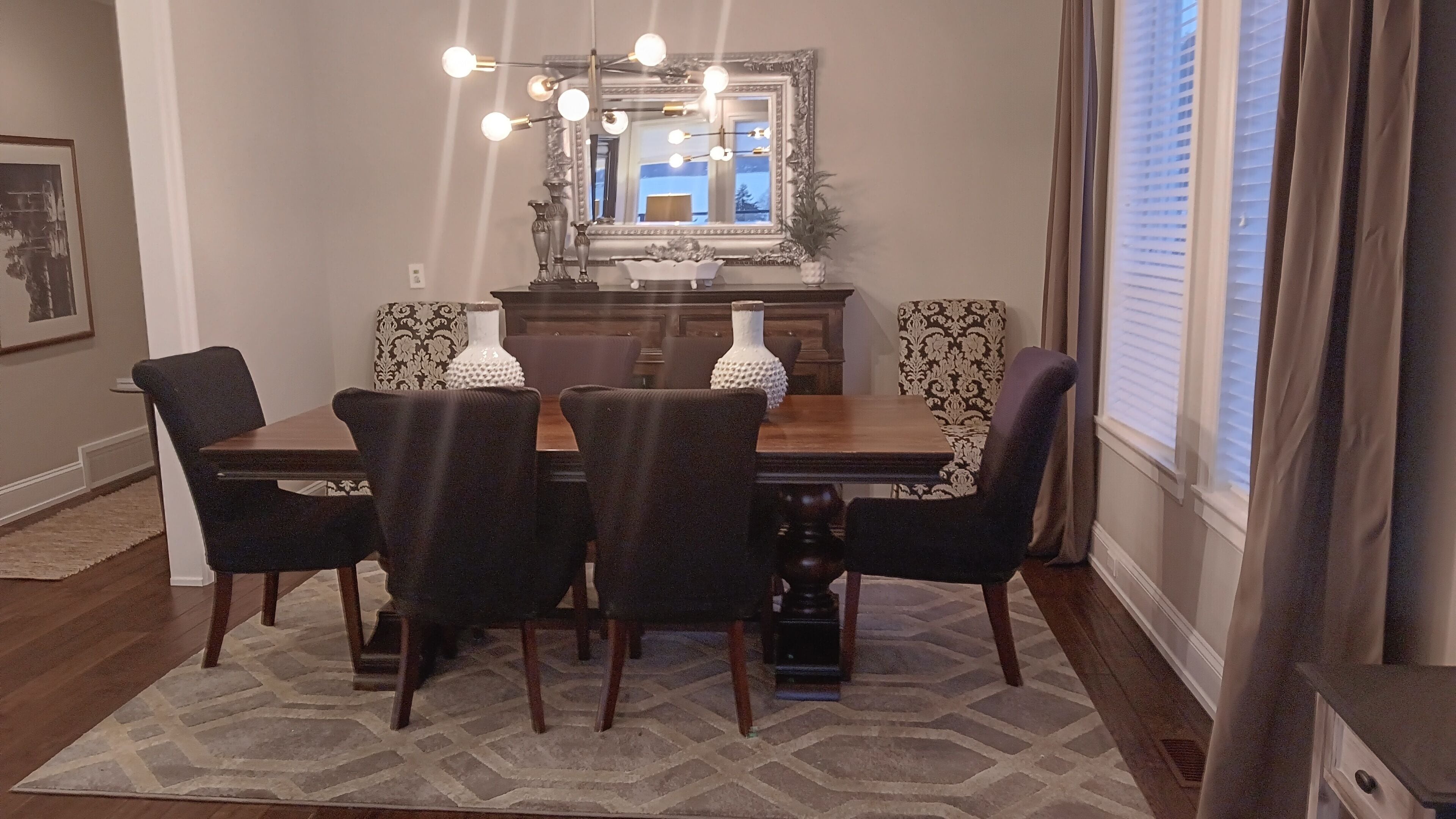 Comedor con una mesa de madera y cuatro sillas, un espejo decorativo y un tapete estampado.