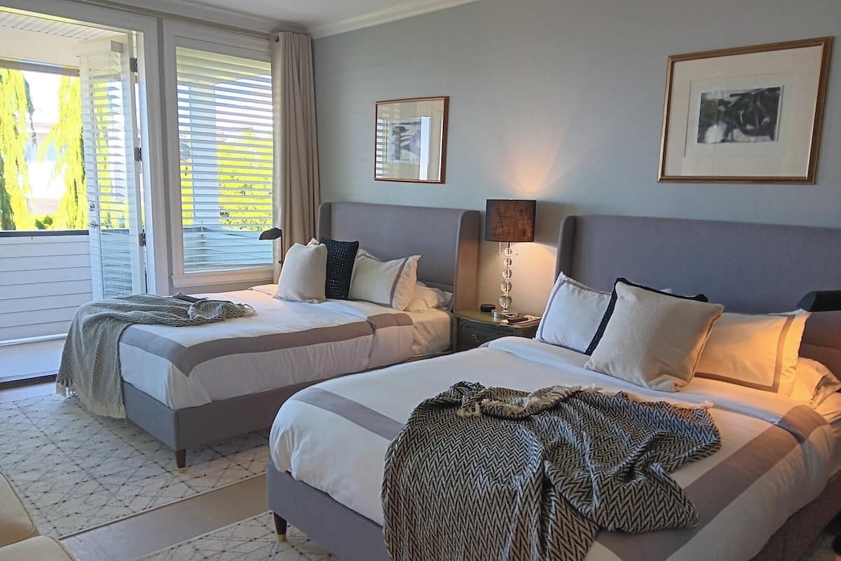 Habitación de hotel con dos camas, un ventanal con persianas, una lámpara y cuadros en las paredes.