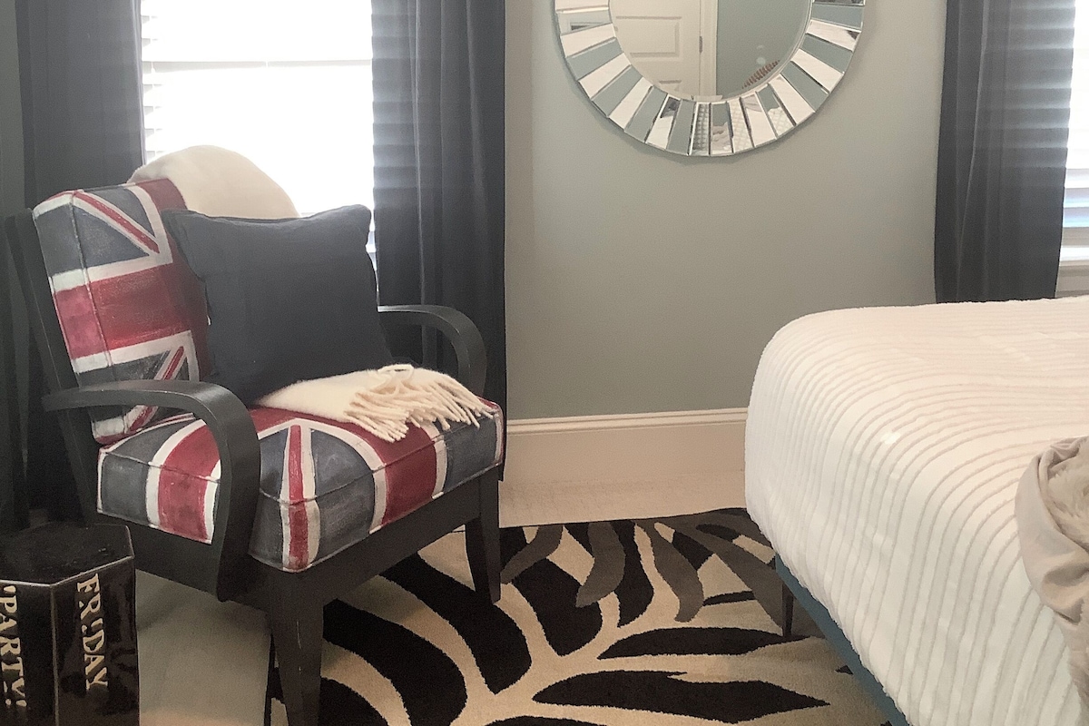 A bedroom with a bed, a chair with a patterned cushion, a round mirror on the wall, and a window with curtains.