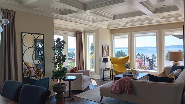 A spacious living room with a large window offering a view of the sea, a cozy sofa, a dining table, and a potted plant. - Sand and Salt Bed & Breakfast (White Rock)