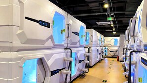 Une rangée de modules blancs, à l’allure futuriste, équipés de panneaux de contrôle et d’espaces d’assise, disposés de manière linéaire le long d’un couloir où des repères directionnels sont indiqués sur le sol.