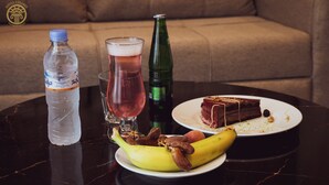 Una mesa con una botella de agua, un vaso de bebida rosa, una botella de refresco verde, un plátano y una porción de pastel en un plato.