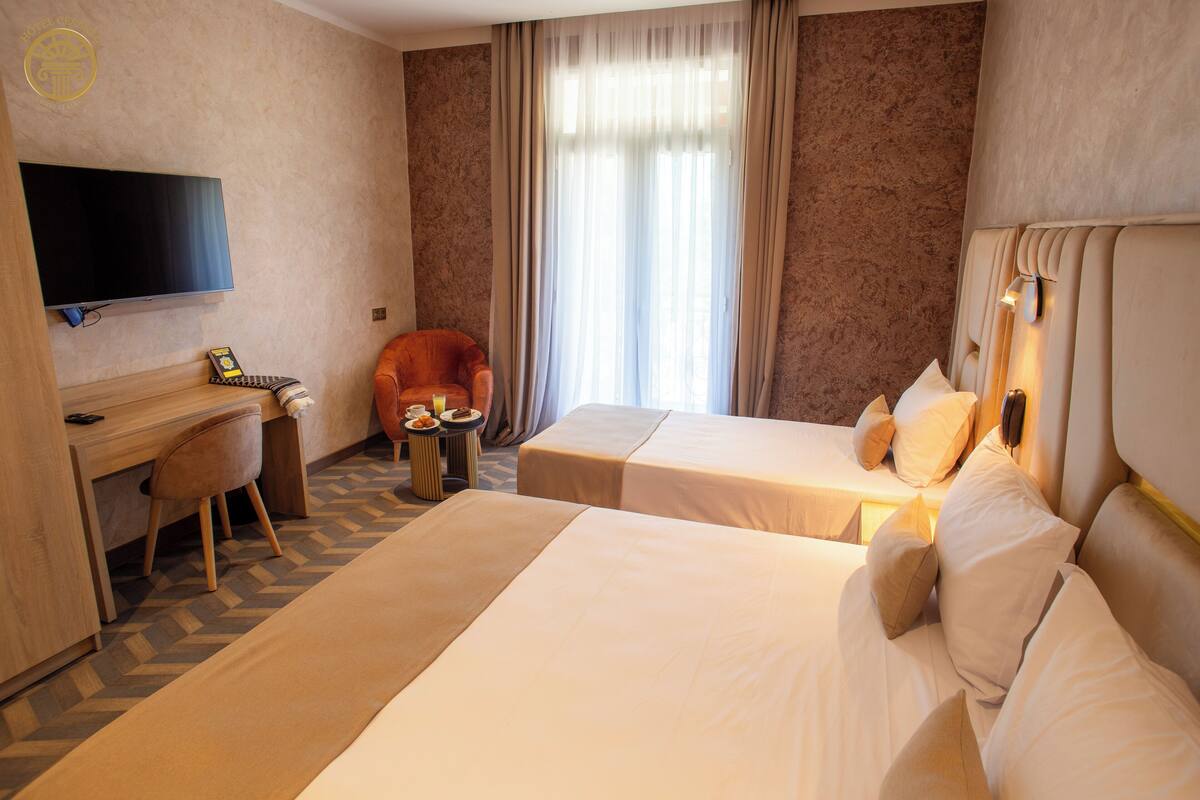 A hotel room with two beds, a desk, a chair, a TV, and a window with curtains.