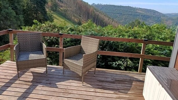 A wooden deck with two wicker chairs and a wooden railing, overlooking a forested valley. - Ferienhaus Meurastein (Meura)