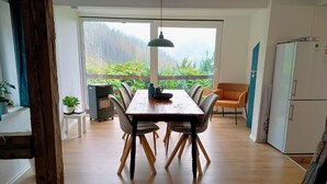 A dining area with a wooden table and chairs, a ceiling light, and a view of greenery through large windows. - Ferienhaus Meurastein (Meura)