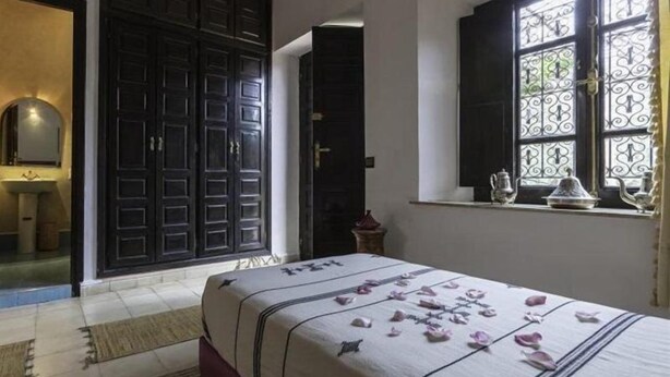 A bedroom with a bed, a window with decorative iron bars, a dark wooden wardrobe, and a bathroom visible through an open door. - Riad Harouak (Souihla)