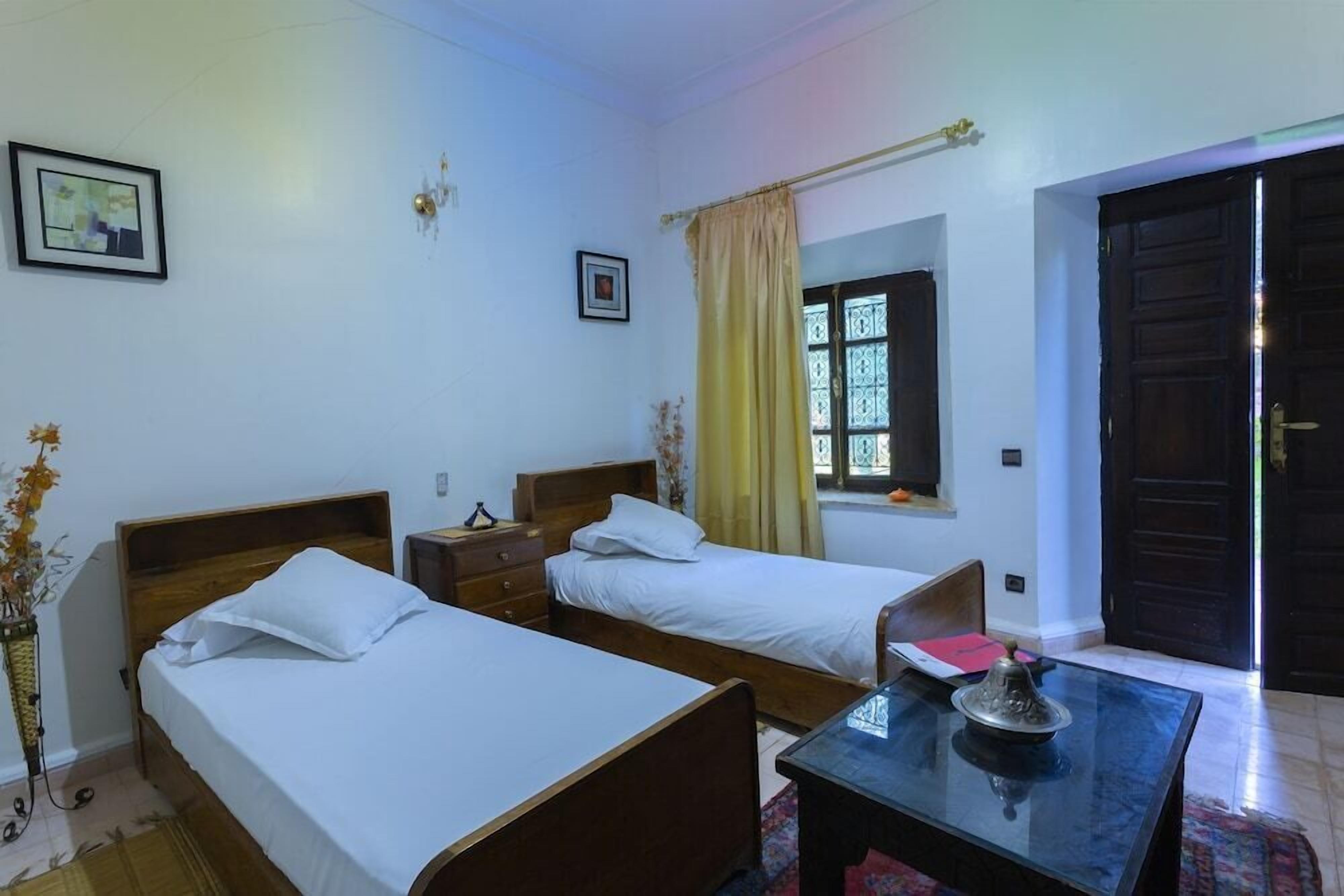 A room with two single beds, a small table with a decorative item, a window with curtains, and a door.