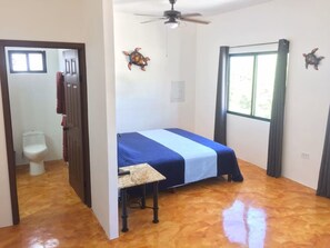 Une pièce comprenant un lit, un ventilateur de plafond, une fenêtre avec des rideaux et une porte donnant sur une salle de bain.