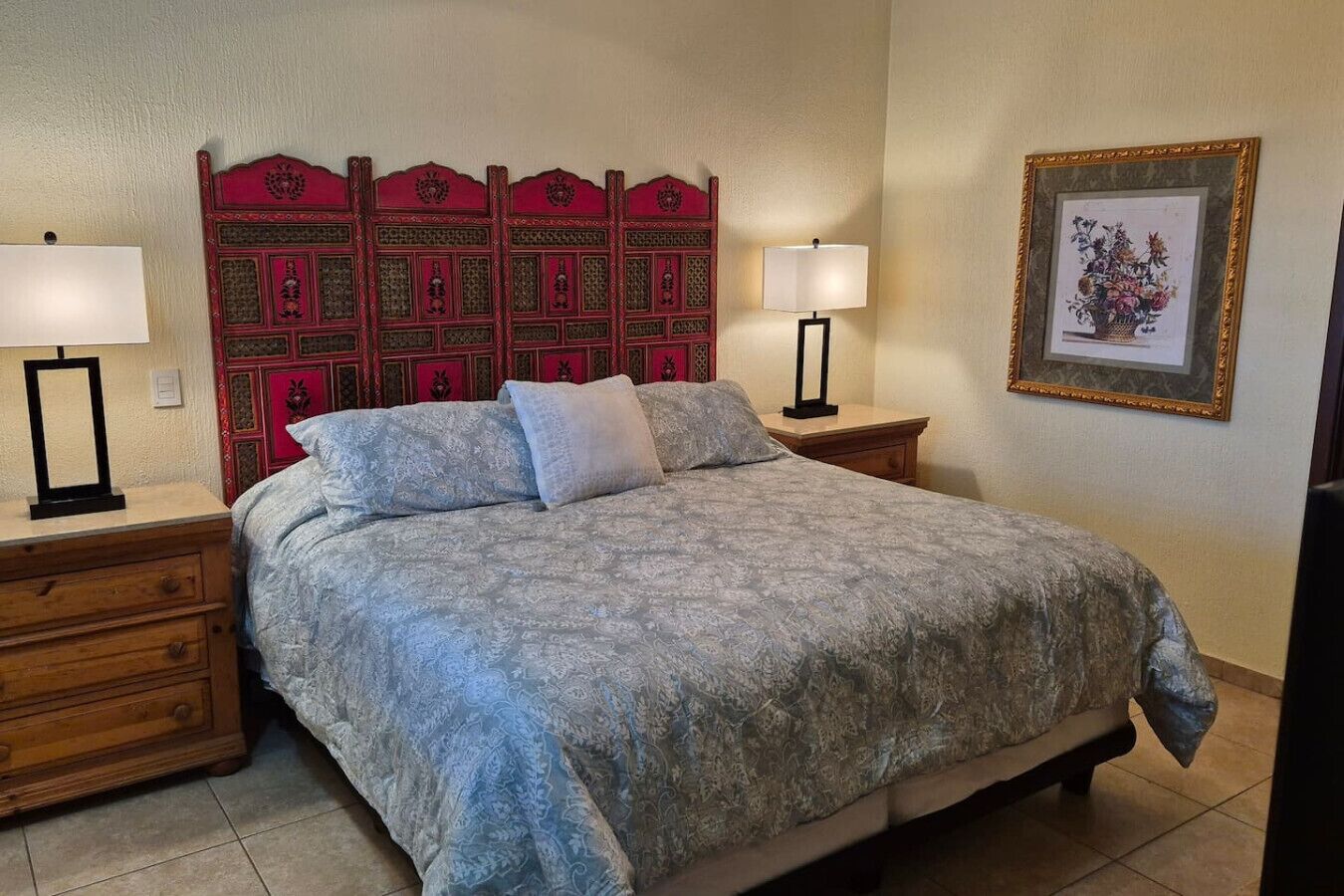 Een slaapkamer met een bed met een geborduurd sprei, twee nachtkastjes met lampen, een houten nachtkastje, een decoratief hoofdbord en een ingelijste foto aan de muur.