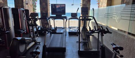 A gym with treadmills, elliptical machines, and weight equipment, overlooking a beach.