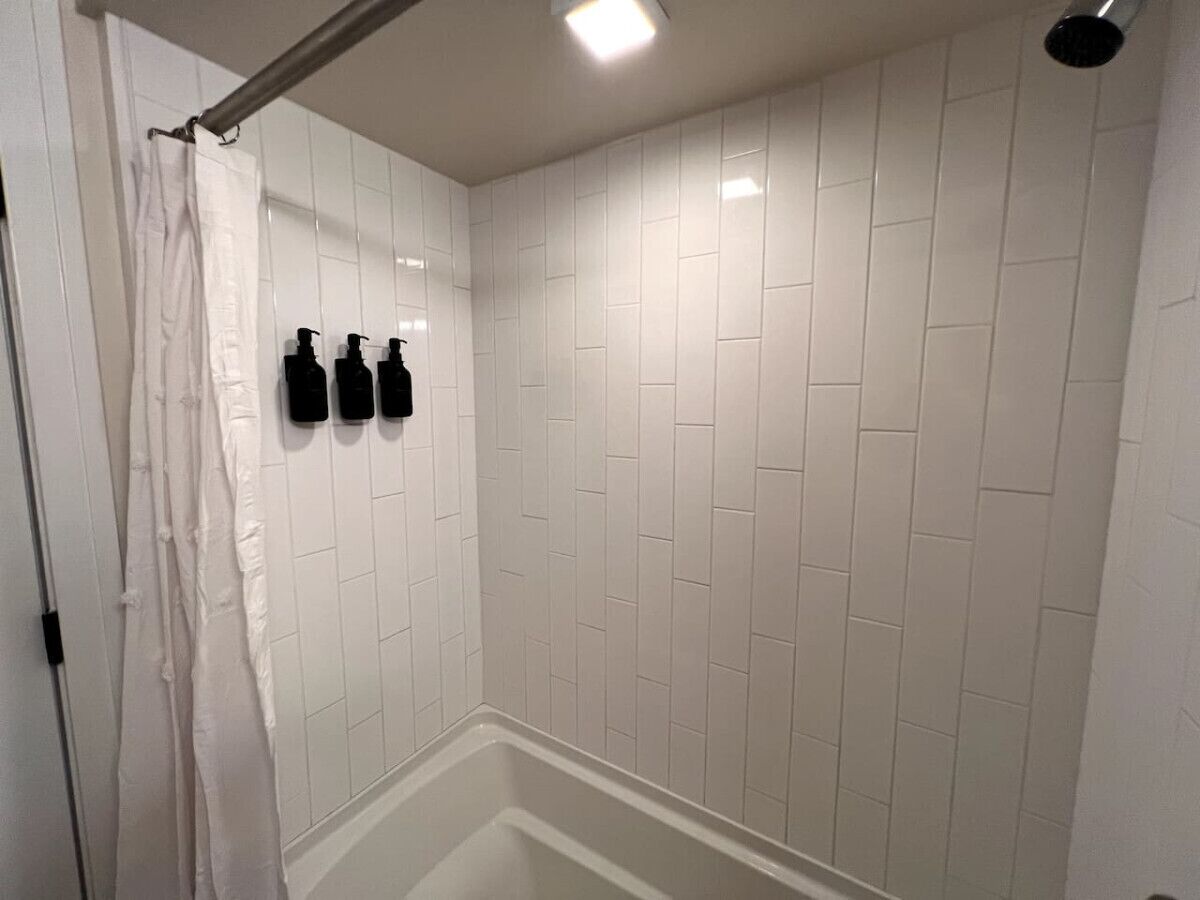Eine Dusche mit weißen Fliesen, einem weißen Vorhang und schwarzen Flaschen an der Wand.