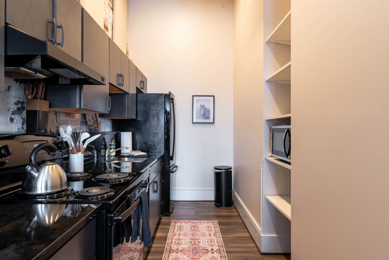A compact kitchen with a stove, oven, and microwave, wooden cabinets, and a small trash bin.