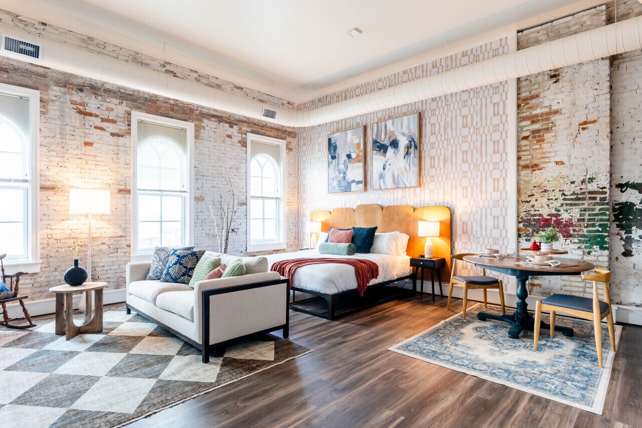 A room with a bed, a sofa, a dining table, and a large window.