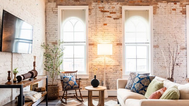 A living room with a brick wall, a TV mounted on the wall, a sofa, a chair, a small table, and a lamp. - Hotel California - KING BED LOFT (Ypsilanti)