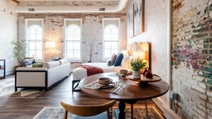 A room with a bed, a sofa, a dining table with chairs, and large windows. - Hotel California - KING BED LOFT (Ypsilanti)