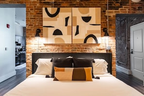 Un dormitorio con pared de ladrillo, cama con ropa de cama blanca y negra, y arte abstracto sobre la cama.