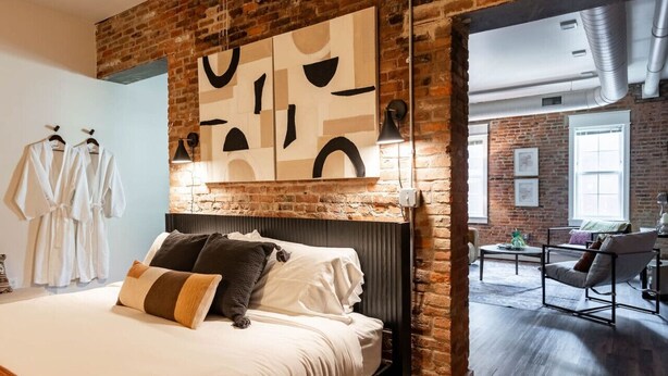 Ein modernes Hotelzimmer mit einer Ziegelwand, einem großen Bett mit Kissen, einem Kopfteil und einem Gemälde. Im Hintergrund befindet sich eine Sitzecke mit Tisch und Stühlen.