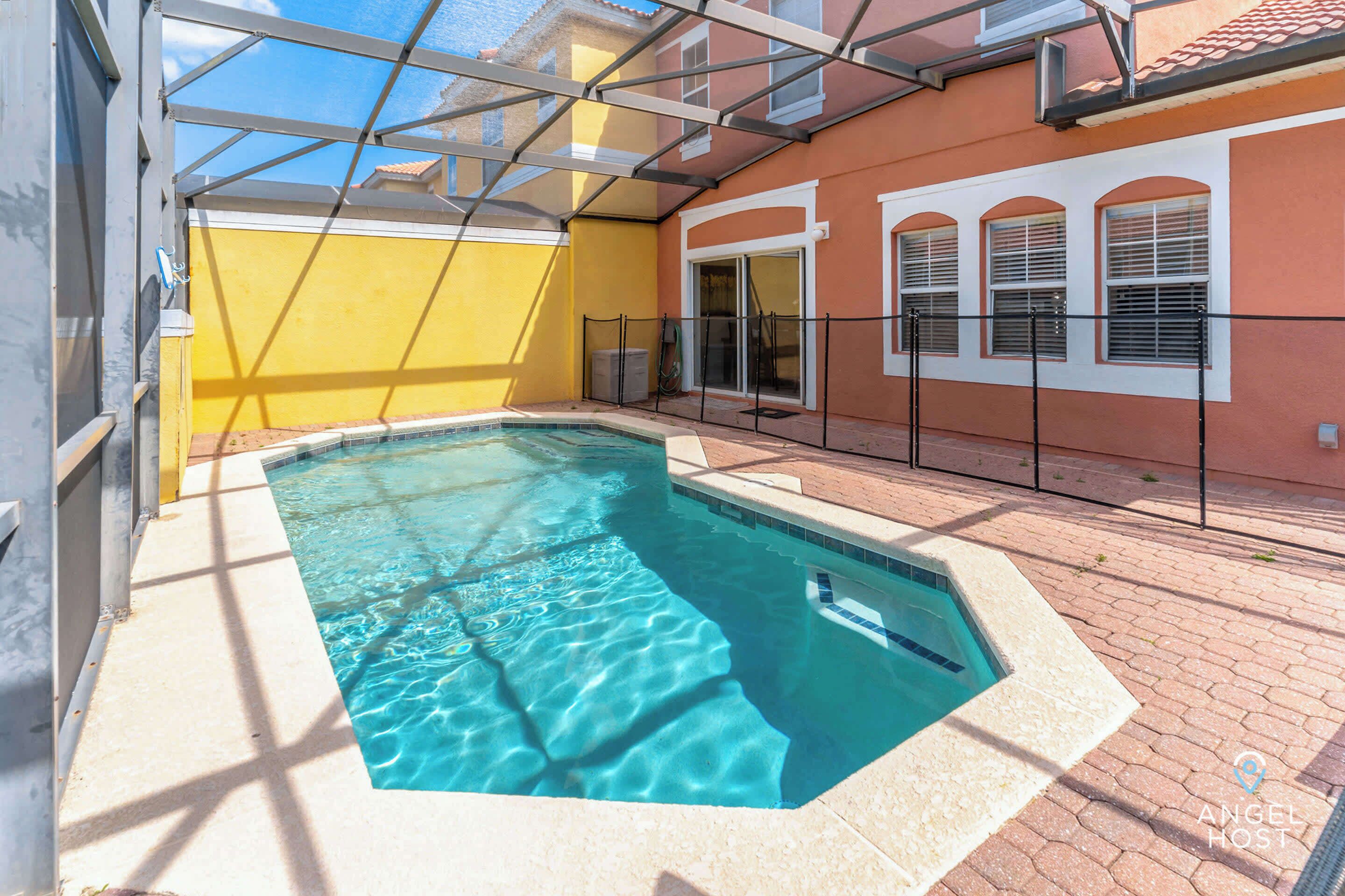 A swimming pool with a glass enclosure, a yellow wall, and a tiled patio area.