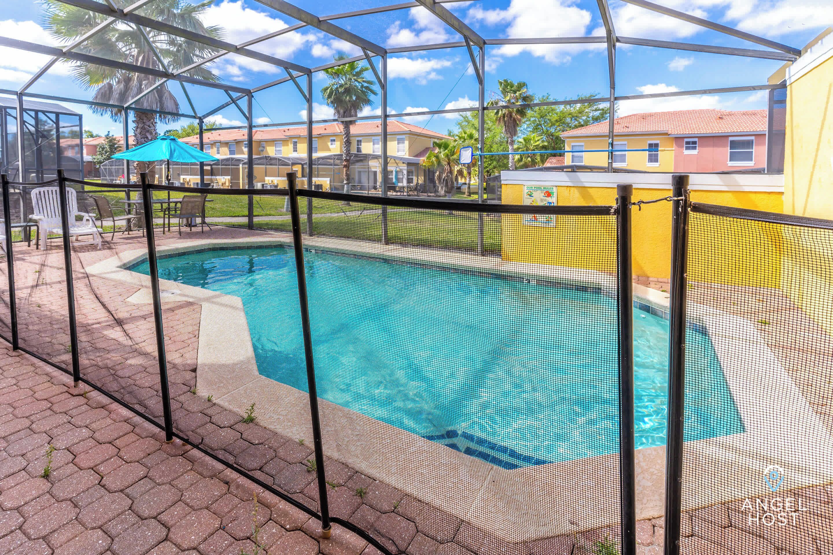 Une piscine entourée d’une clôture de sécurité, bordée d’une terrasse couverte avec des chaises et un parasol bleu.