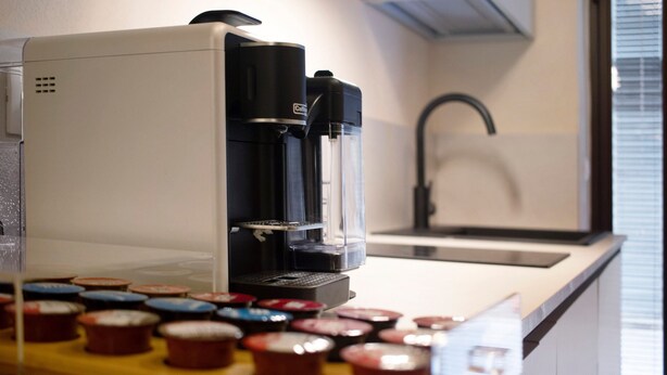 Une machine à café avec une verseuse et un bac de récupération, un évier et une fenêtre avec des stores.