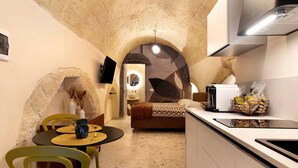 Eine kompakte Küche mit einem Steingewölbe, ein Essbereich mit Stühlen, ein Bett und ein an der Wand befestigter Fernseher.