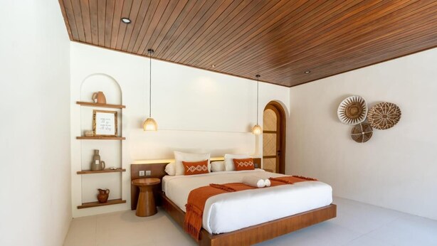 A modern bedroom with a wooden ceiling, a large bed with white bedding and orange accents, a bedside table with a lamp, and decorative wall art. - Jimbaran Bay Villas (Kedonganan)