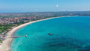 An aerial view of a coastal city with a long sandy beach, a pier with boats, and a densely populated urban area in the background. - Jimbaran Bay Villas (Kedonganan)