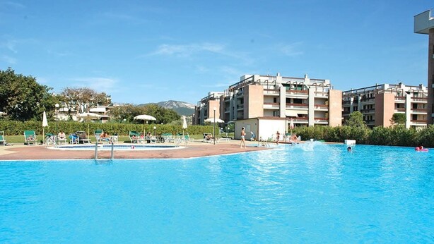 A swimming pool with lounge chairs and umbrellas, surrounded by a building with multiple balconies and a mountain in the background. - 5 bed places with swimming pool in the area Coop (Toscana)