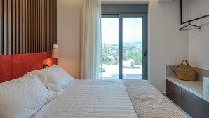A hotel room with a bed, a view of the city through a large window, a hanging light fixture, and a small table with a basket.