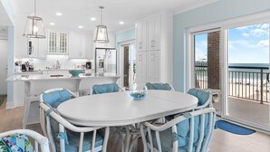 A modern kitchen with white cabinets, a dining area with a round table and blue chairs, and a balcony with a view of the beach. - Beachfront Bliss: 3 Bdrm 1st Flr (Pawleys Island)