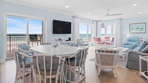 A spacious living area with a round table and chairs, a flat-screen TV, and a view of the ocean through large windows. - Beachfront Bliss: 3 Bdrm 1st Flr (Pawleys Island)