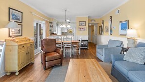 Una sala de estar acogedora con un sillón de cuero marrón, un sofá azul y una mesa de centro de madera. Se observa un comedor con mesa y sillas, y una cocina al fondo.