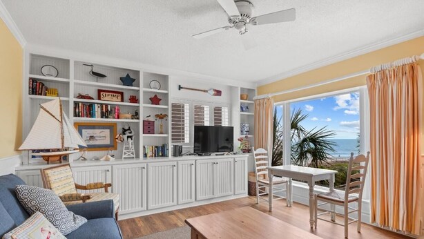 Un salón con un sofá, una mesa de madera y un televisor. Se ve una estantería con varios objetos y una ventana con cortinas.