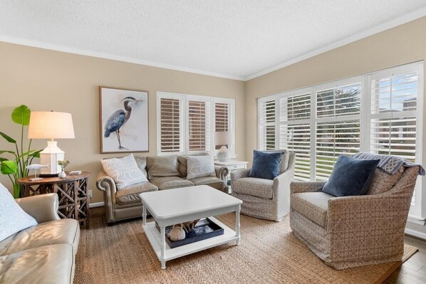 Ein Wohnzimmer mit einem beige-farbenen Sofa, einem gemusterten Sessel, einem weißen Couchtisch und einer Lampe. Zwei Fenster mit Fensterläden und ein gerahmtes Bild von einem Vogel sind an der Wand zu sehen.