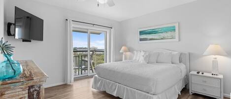 Quarto com cama, ventilador de teto, televisão fixada na parede, mesa de cabeceira com luminária e janela com vista para o exterior.