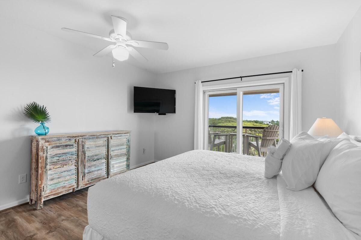 A bedroom with a large bed, a ceiling fan, a TV mounted on the wall, and a view of the outdoors through a sliding door.