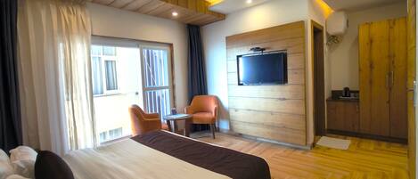 A modern hotel room with a large bed, a TV mounted on a wooden wall, a small table with a chair, and a kitchenette area.