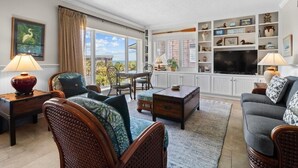 A spacious living room with a large window, wicker furniture, a television, and a bookshelf.