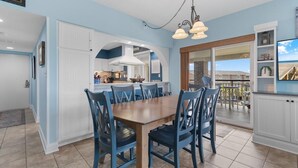 Un coin repas avec une table en bois et des chaises bleues, une cuisine en arrière-plan et une vue sur un balcon avec des sièges d’extérieur.